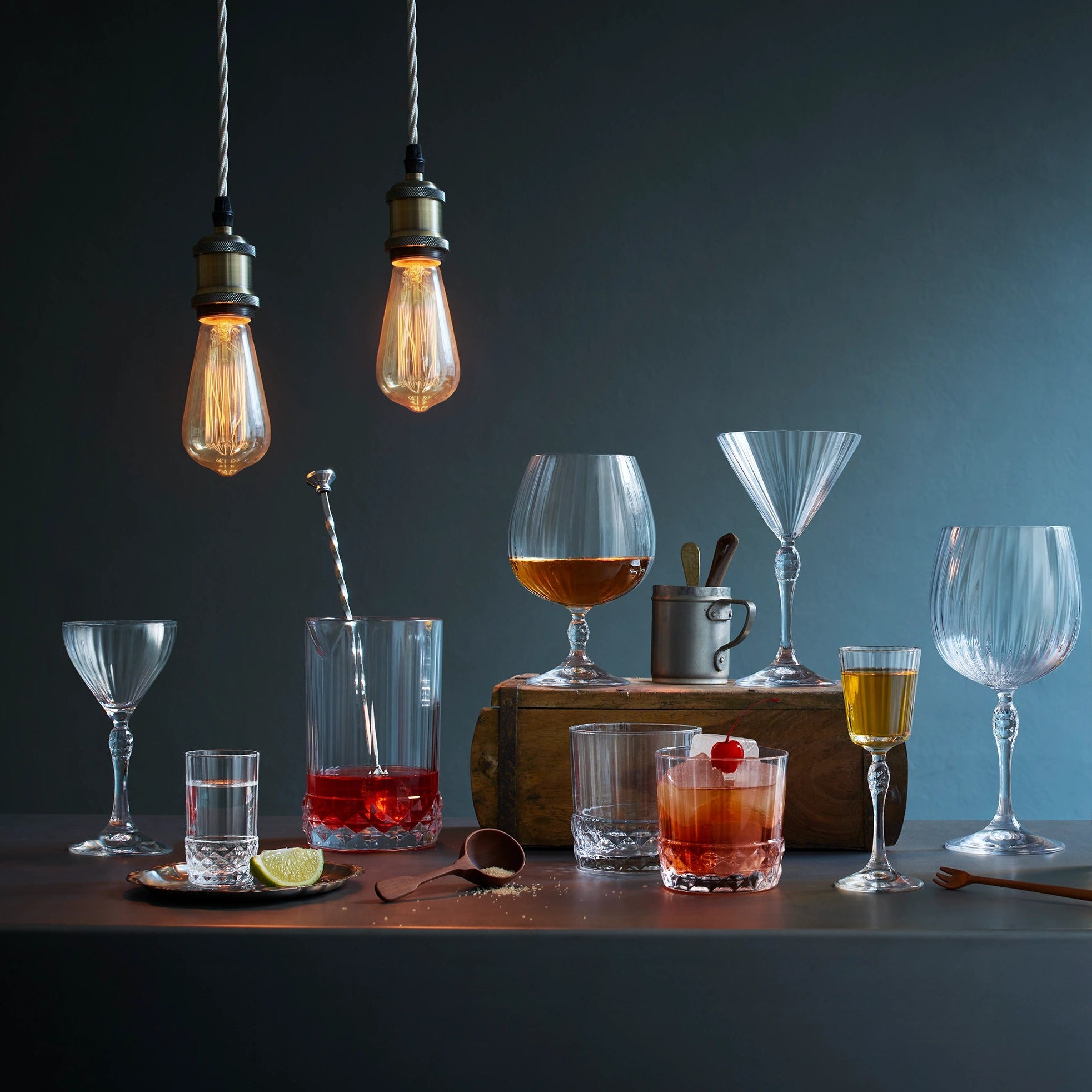 A bar-style still life with multiple clear drinking glasses and cocktail glassware arranged on a dark tabletop against a blue-gray background. Two hanging Edison bulbs illuminate the scene, with drinks, straws, a jigger, flowers in a vase, and wooden blocks used as styling props.