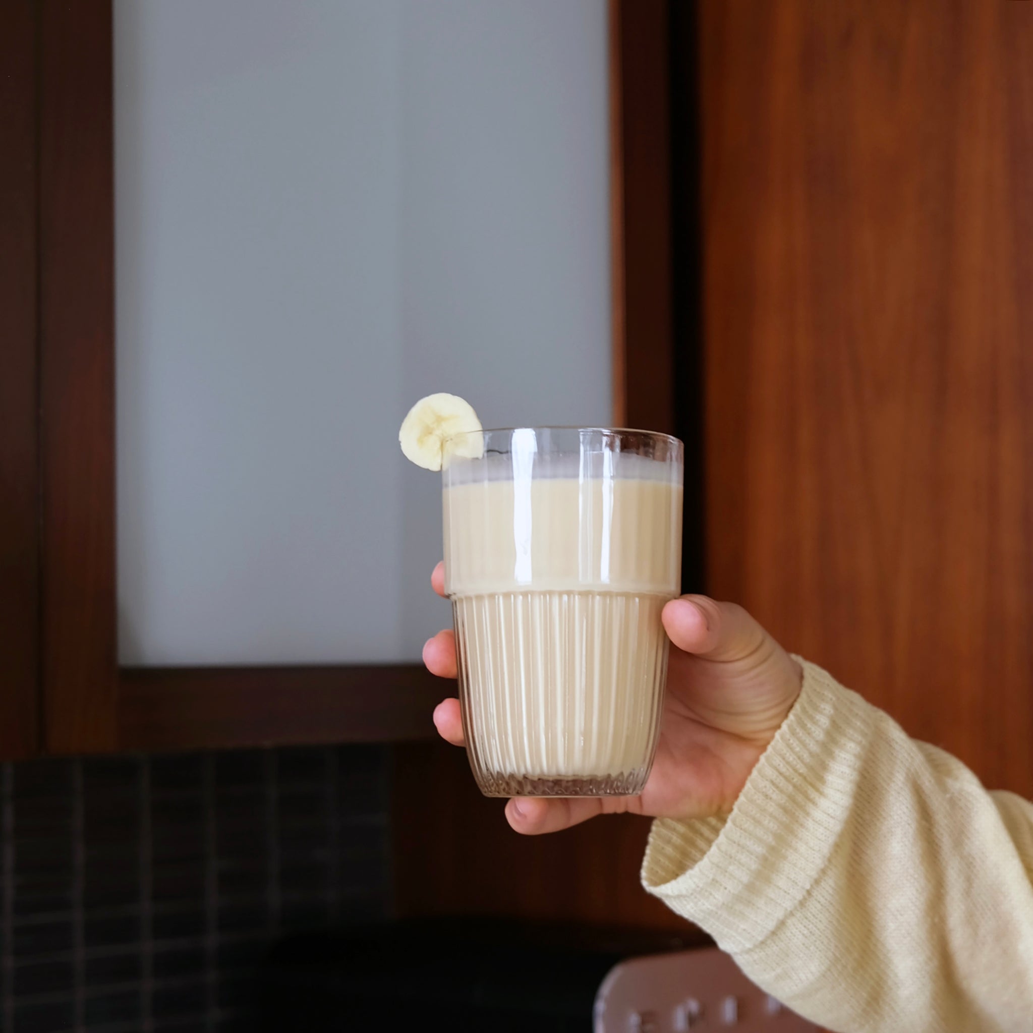 Hand holding a glass of milkshake with a banana slice against a blurred indoor background