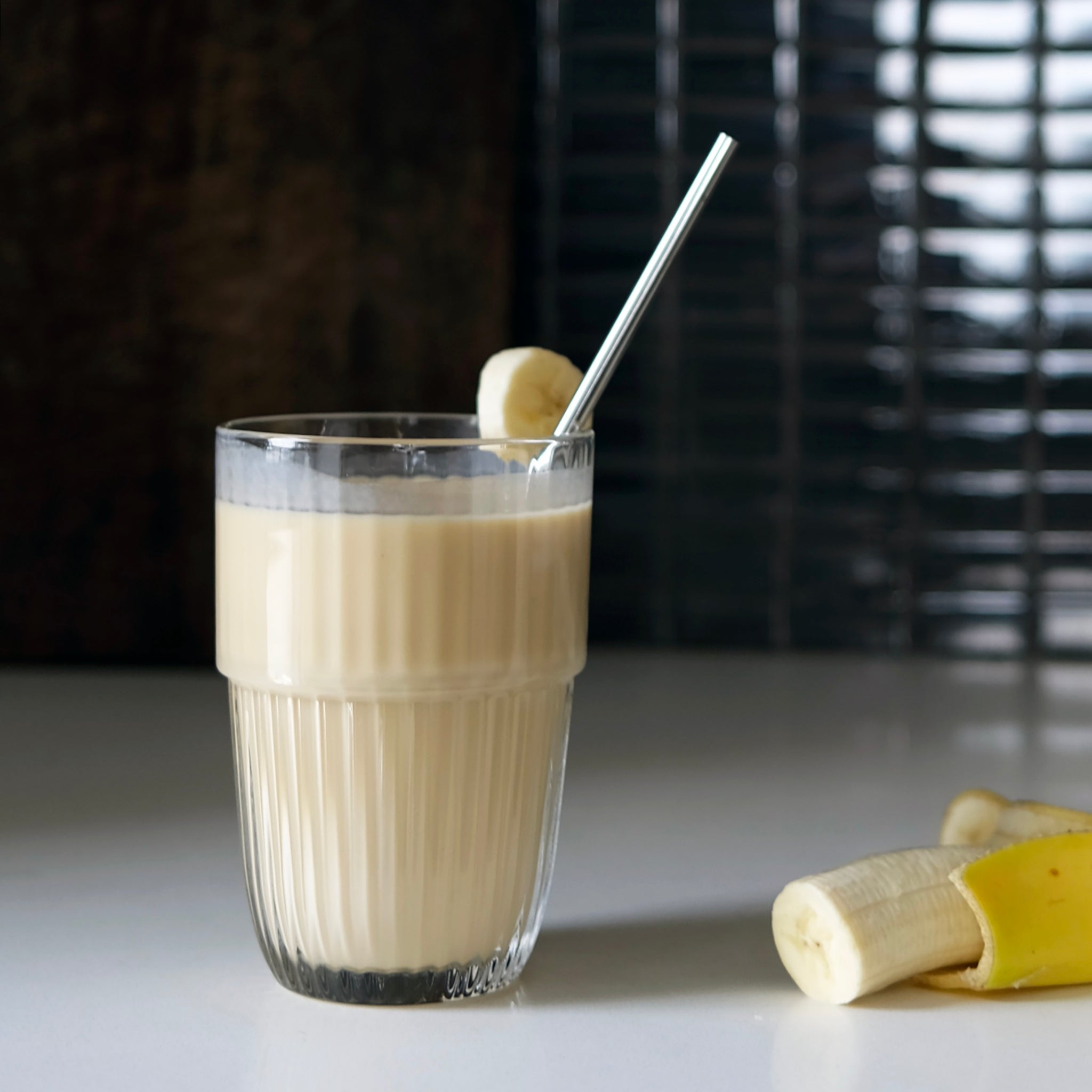 Glass of banana smoothie with a straw on a white surface, with bananas in the background.