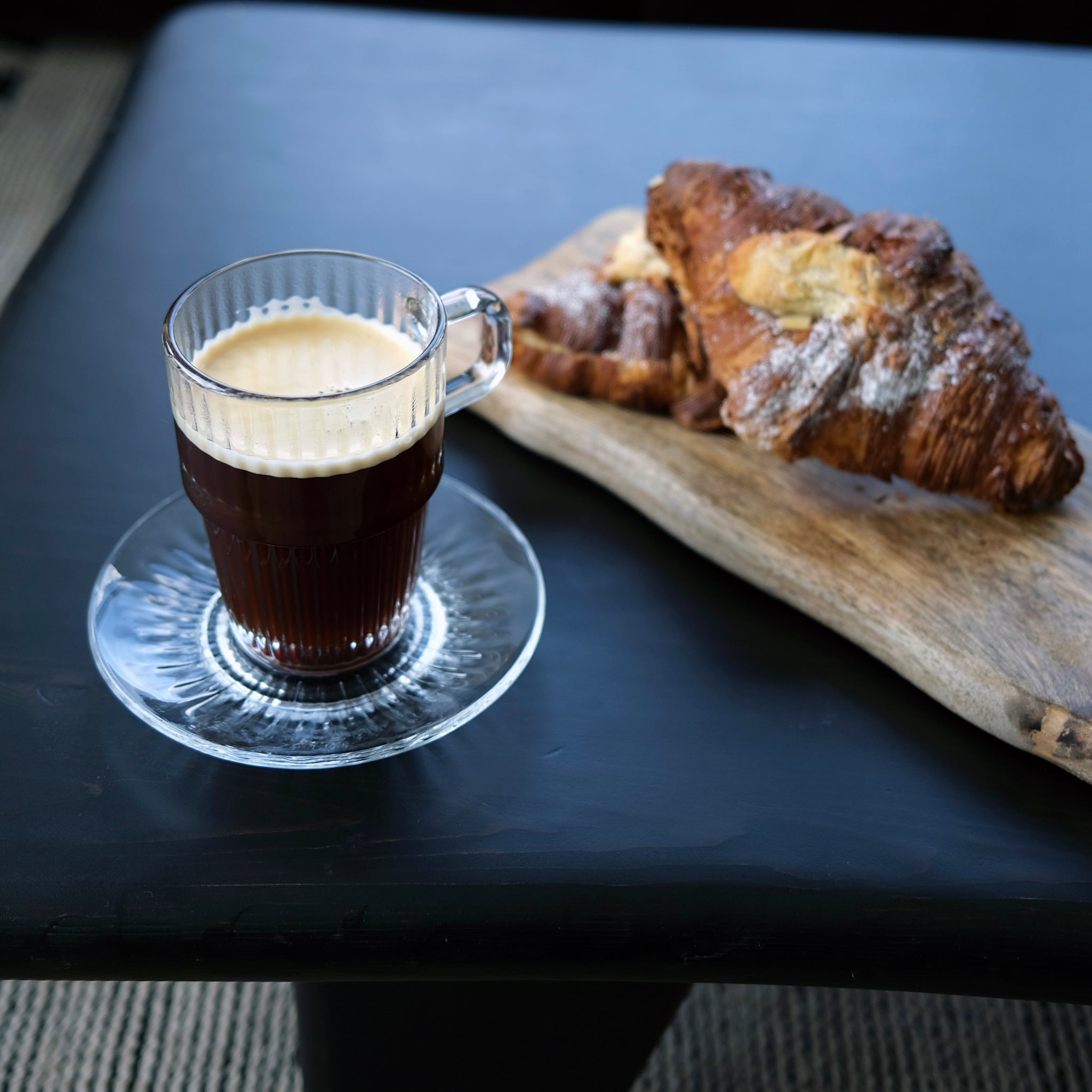 Glass of coffee with a saucer on a dark surface with a wooden board of pastries.