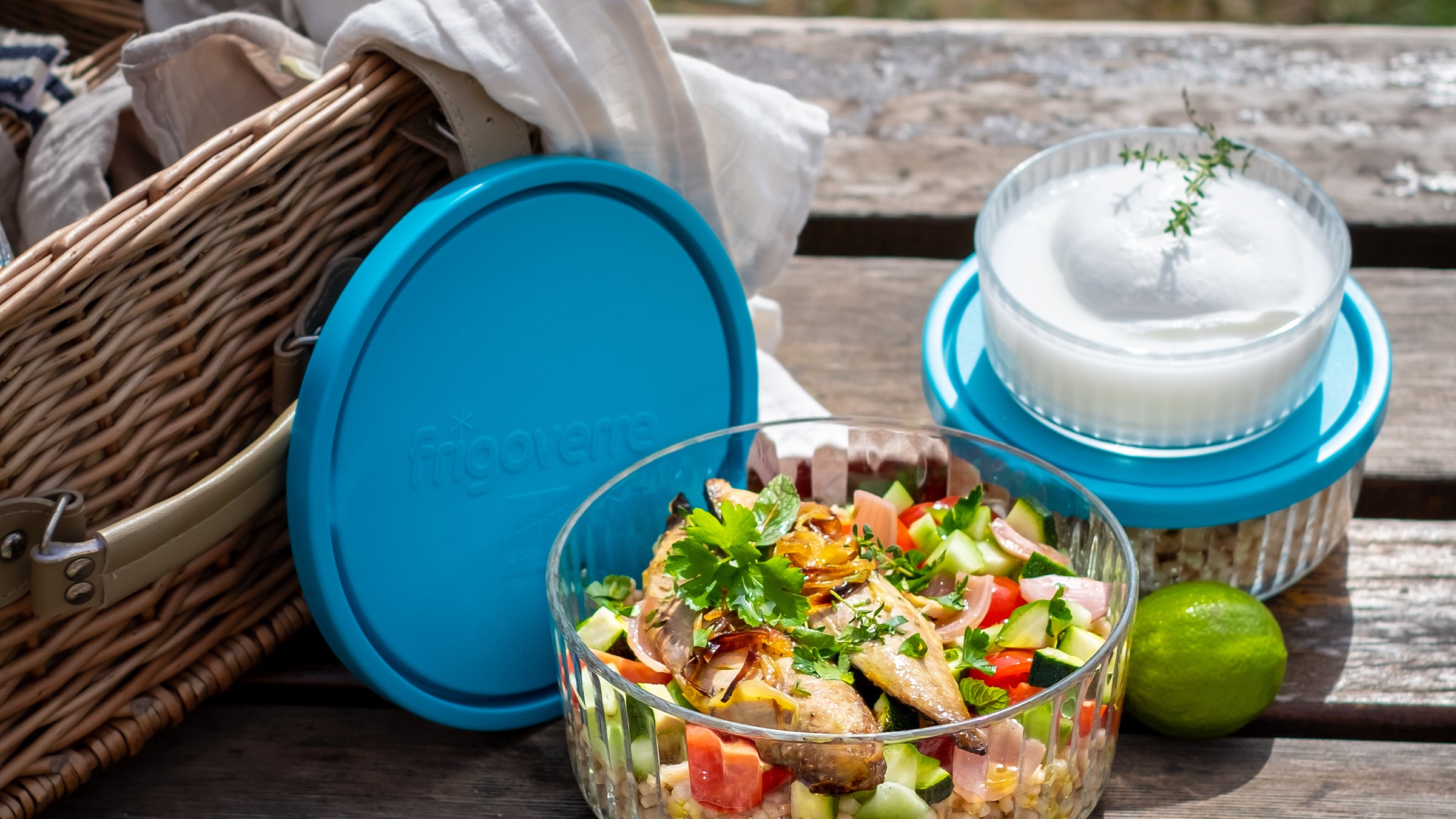 Picnic setup with Frigoverre Brillante salad bowl, blue container lids, and picnic basket on wooden table.