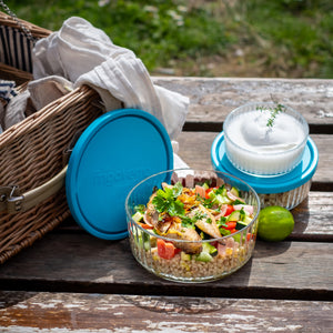 Picnic setup with Frigoverre Brillante salad bowl, blue container lids, and picnic basket on wooden table.