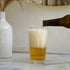 Sestriere Beer glass with beer being poured next to a white vase with dried plants on a marble surface.