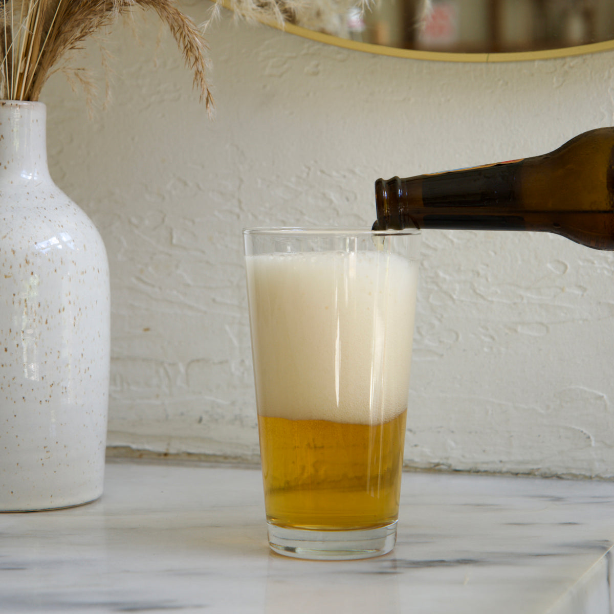 Sestriere Beer glass with beer being poured next to a white vase with dried plants on a marble surface.